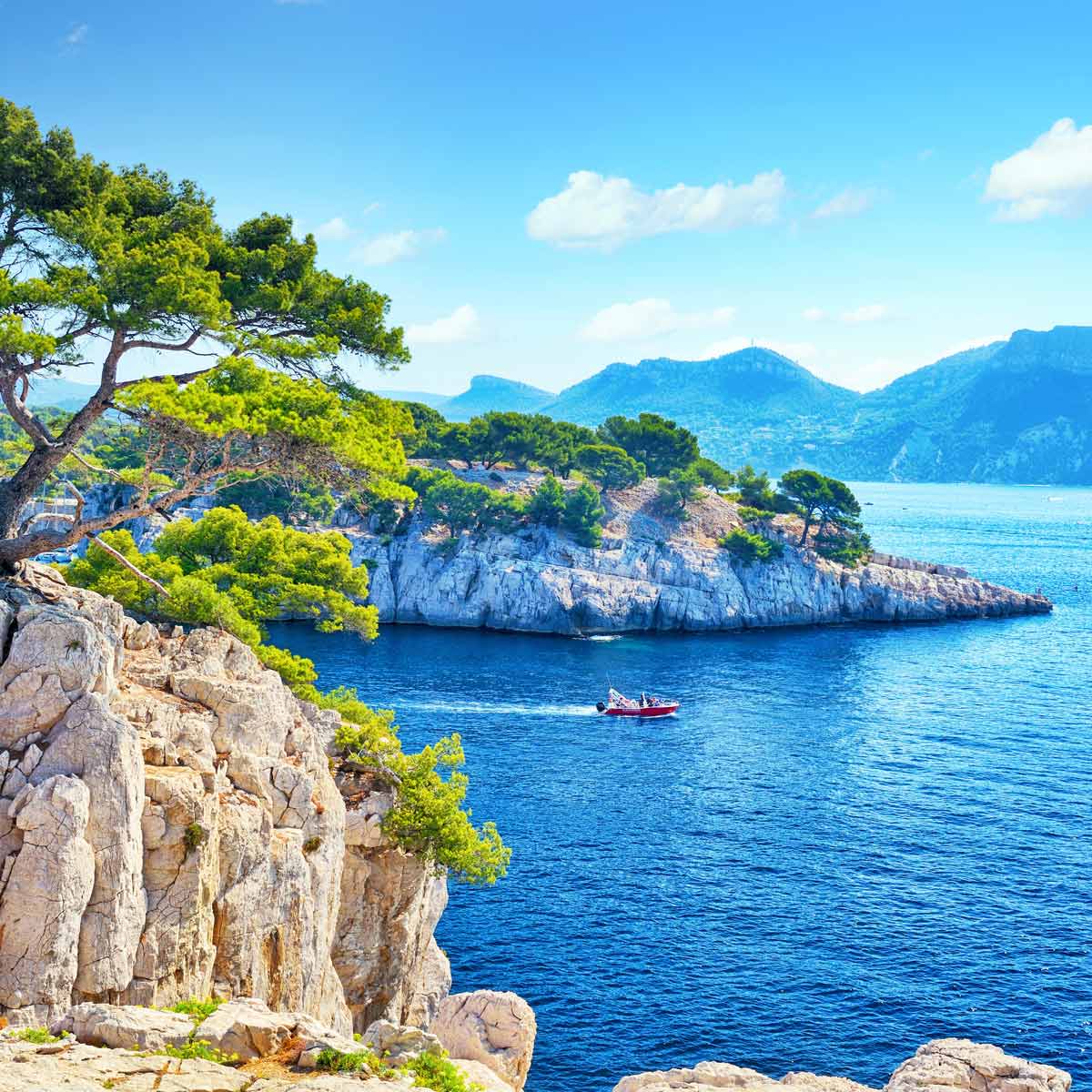 A red boat sails on the deep blue waters in front of the rocky point of Port-Miou.