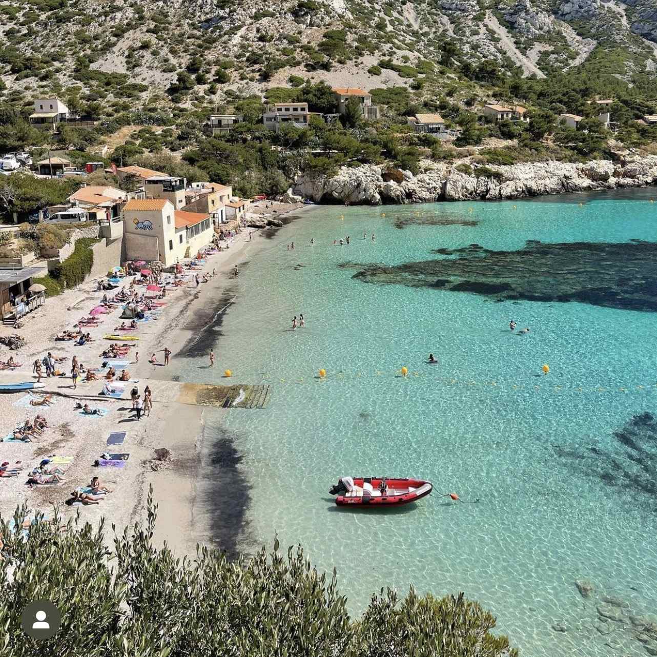 High-angle bird's-eye view of the pebble beach, traditional stone cottages, and clear, turquoise waters of the Sormiou calanque in Marseille.